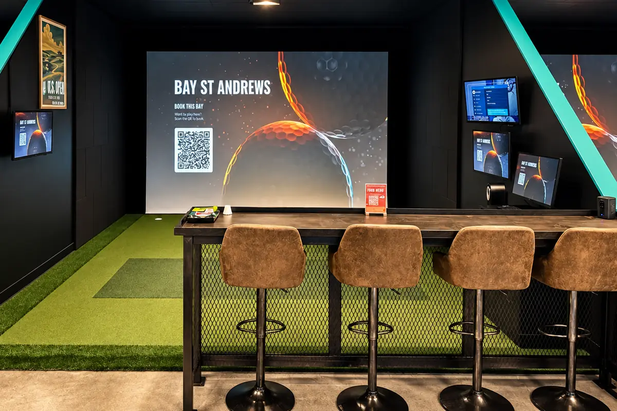 Bay St Andrews: a big screen over a turf hitting area, with a bar-height table and four stools along the right.