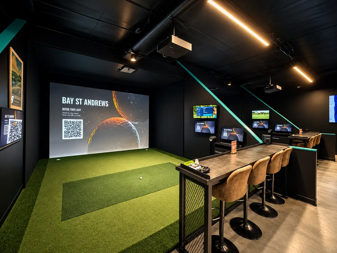 A bay seen from the side, with a lit screen, a turf hitting mat, teal accents on black walls and a bar table with stools. More bays continue beyond it.