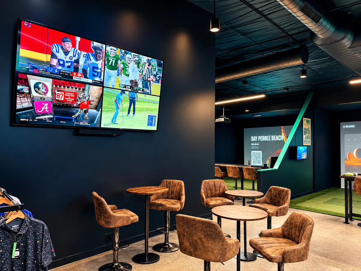 The lounge: a big wall-mounted TV split into four sports feeds, above leather bar stools and round high-top tables, with a bay glowing in the background.
