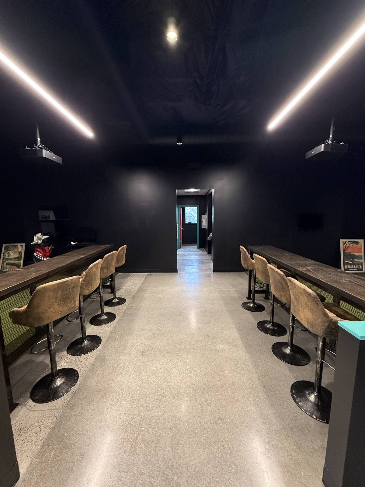 The polished concrete aisle running down the centre of the studio, with long counter rails and rows of suede stools on both sides, framed golf course prints on the end walls, and a lit doorway at the far end.