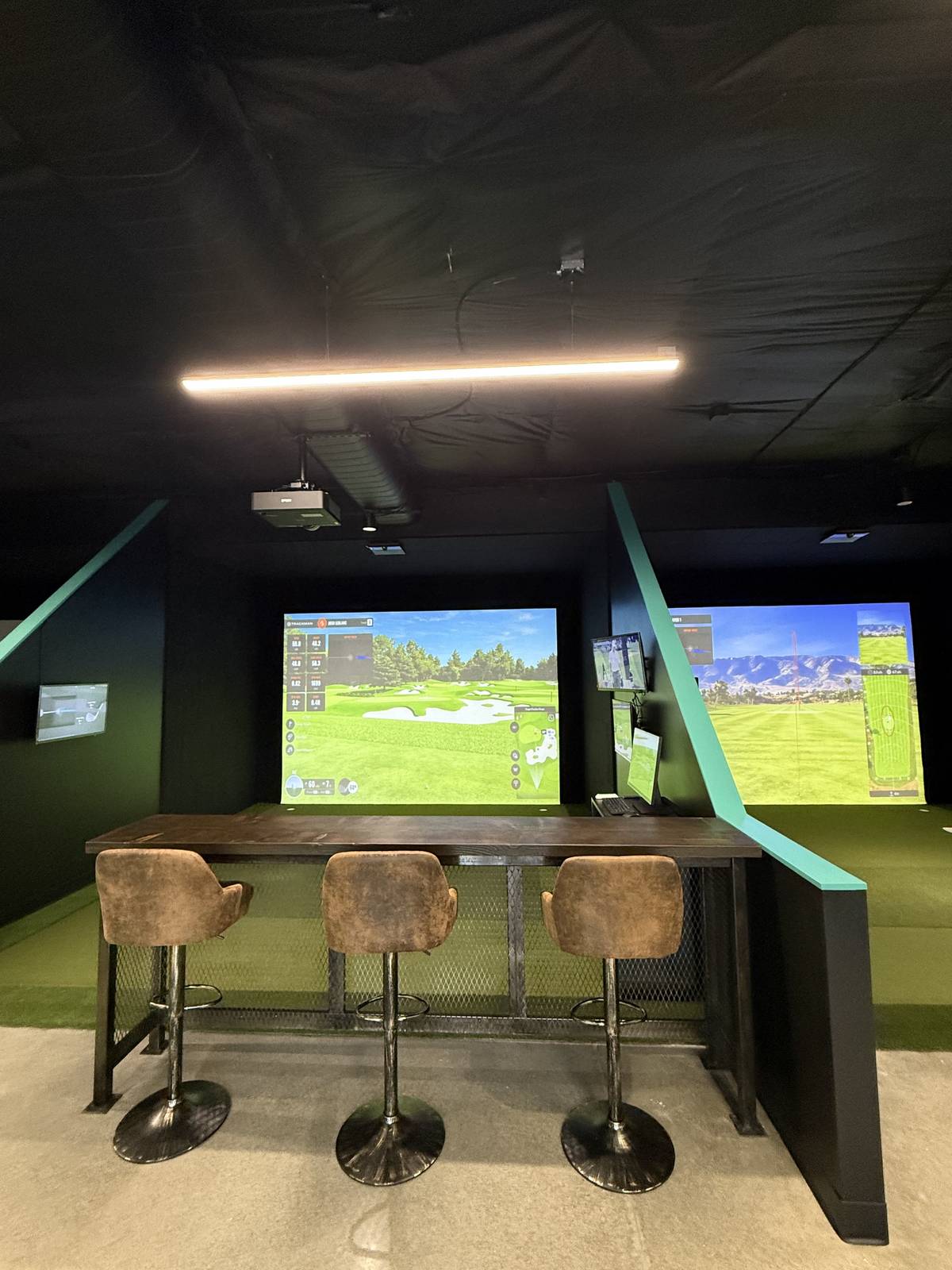 Three suede bar stools tucked under a dark wood high top counter, set between two hitting bays whose screens show different courses. A teal painted divider angles up on each side and a warm strip light runs across the black ceiling.