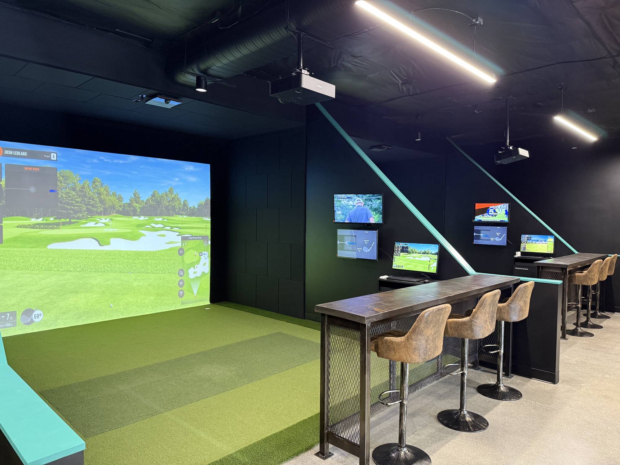 Lit indoor hitting bay with a golf course projected across a wall-width screen, striped green turf underfoot, and a dark wood high top counter with three suede stools facing it. A second bay and its console stand behind, under teal accent stripes.