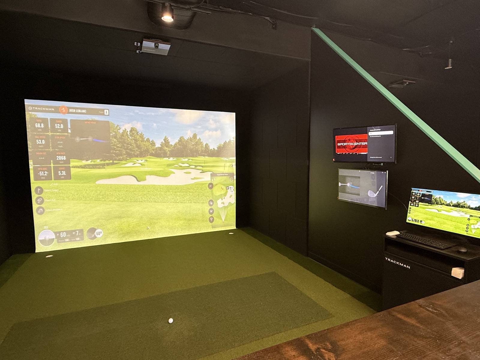 A hitting bay with a par three hole projected on the screen and a shot readout down the left side showing carry distance, ball speed and spin rate. A ball sits on the turf and the bay console with keyboard and monitor stands to the right.